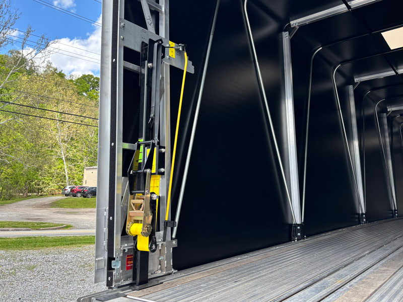 Vango21-rear-post Open trailer interior with a yellow ratchet strap and pulley near the doorway; ribbed metal floor, black walls, outdoors with trees and parked cars in the background.